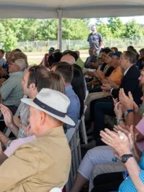 A group of people seated under a tent, clapping during an outdoor event. The audience includes men and women of various ages, some wearing hats. Trees and a fence are visible in the background.