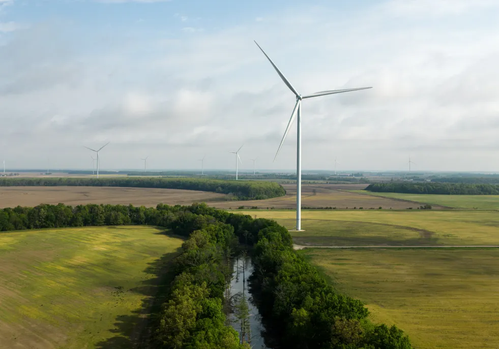 A landscape with several wind turbines scattered across open fields. A large wind turbine is prominent in the foreground, with a narrow stream bordered by trees running through the fields. The sky is partly cloudy, and the horizon is visible in the distance.