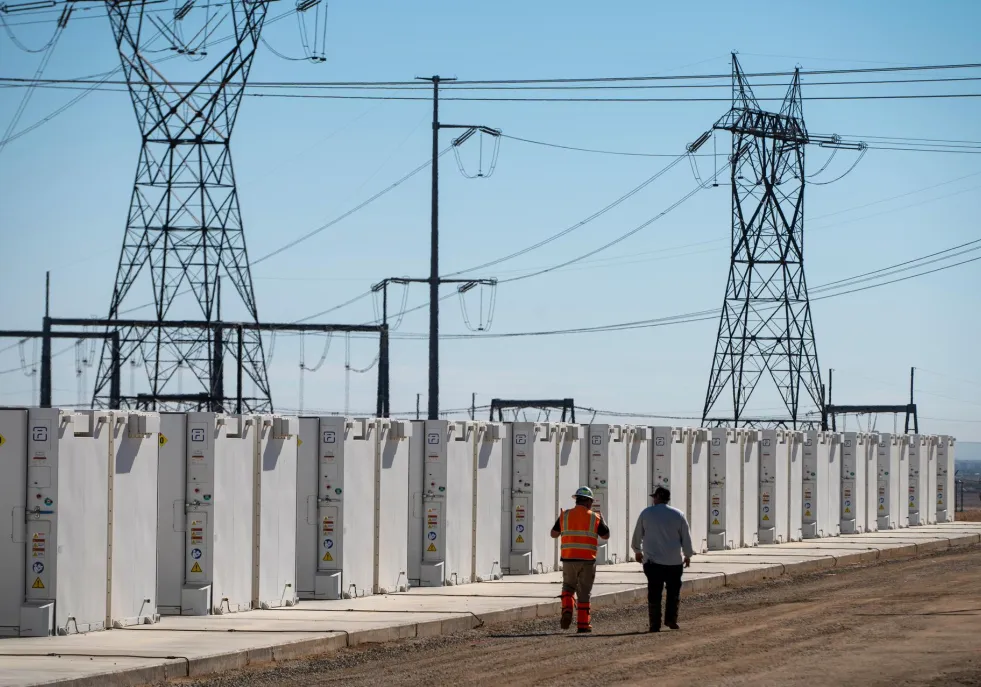Two workers walk alongside a row of white industrial battery storage units in a power facility. High voltage transmission towers and power lines are visible in the background under a clear blue sky.