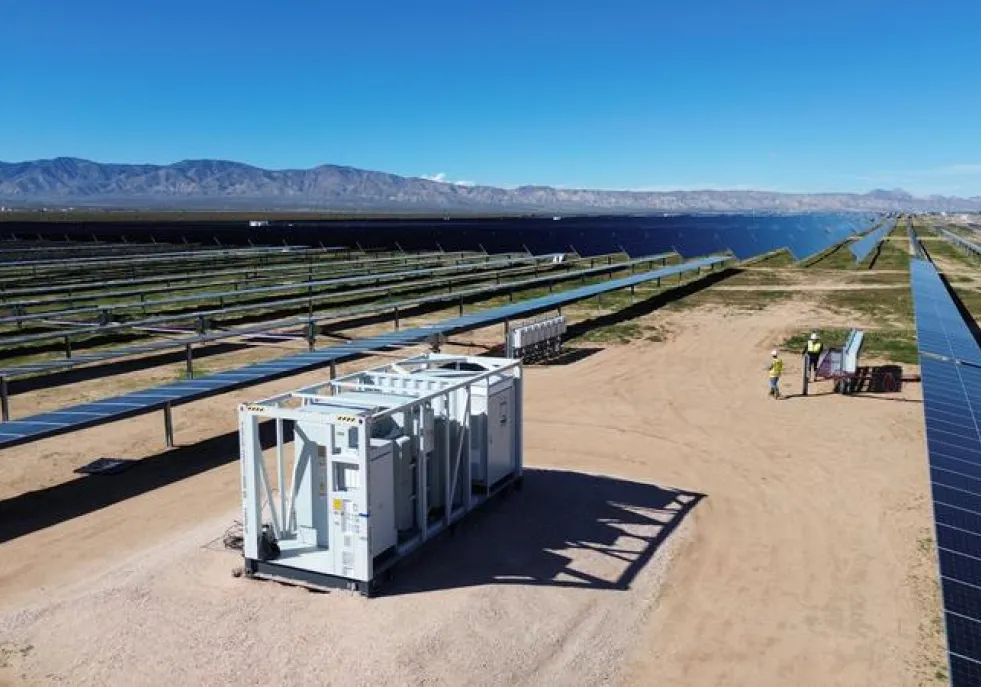 A large solar farm with rows of solar panels stretching into the distance under a clear blue sky. In the foreground, a white utility container is placed on a dirt path. Two people wearing safety gear are near a small table with equipment. Mountains are visible in the background.