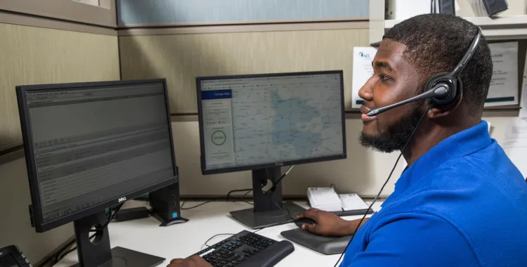 A man wearing a blue polo shirt and a headset sits at a desk in an office cubicle, working on a computer with two monitors. One monitor displays a map, while the other shows a list of data. A telephone is placed on the desk beside the monitors.