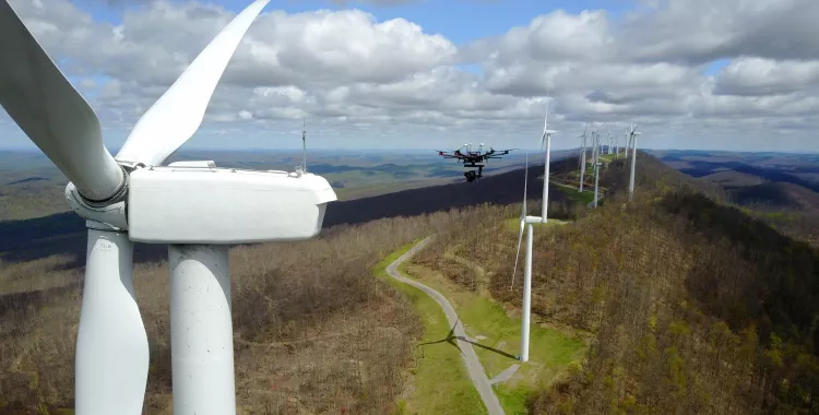 Aerial view of a wind farm with several wind turbines on a hilly landscape. A drone is flying near the turbines under a partly cloudy sky.