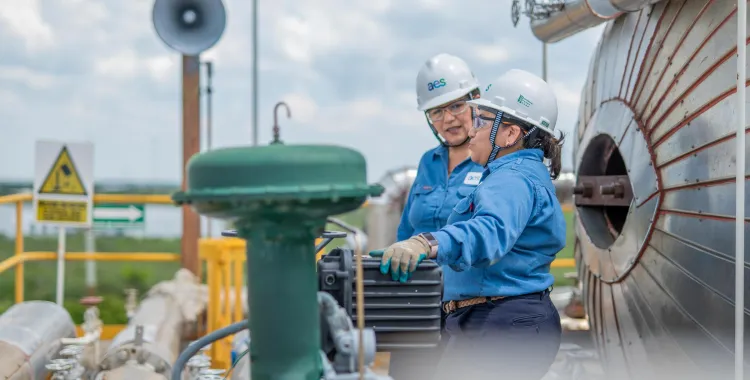 Two workers wearing blue uniforms and white helmets with AES logos are inspecting equipment at an industrial site. They are surrounded by metal pipes and machinery. A warning sign and a loudspeaker are visible in the background.