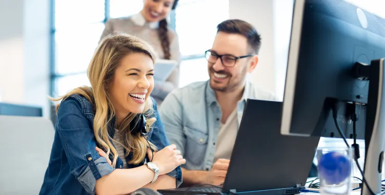 Three people in an office setting, two sitting at a desk with a computer, smiling and laughing. A woman with long blonde hair is in the foreground, and a man with glasses is beside her. A third person stands behind them, smiling and holding a tablet.