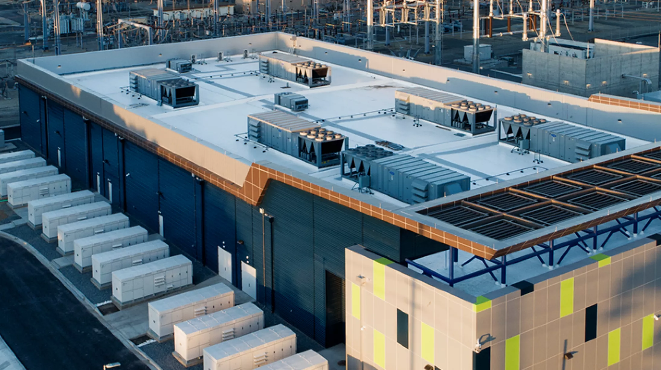 Aerial view of a large industrial facility with a flat roof, featuring multiple HVAC units. The building has a modern design with blue and green accents. Surrounding the facility are several smaller rectangular structures and a network of power lines and equipment, indicating an energy-related site.
