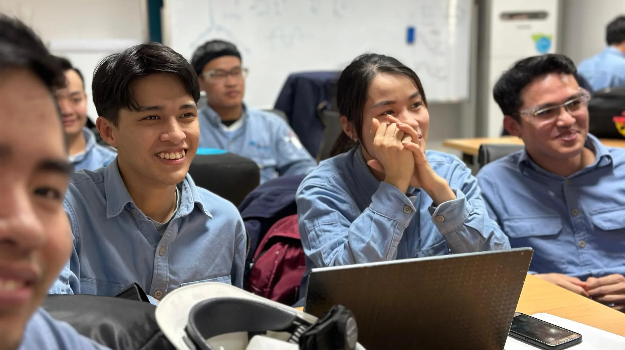 A group of young adults in a classroom setting, wearing blue uniforms, are sitting at desks. They appear engaged and are smiling, with one person resting their chin on their hands. A laptop, a hard hat, and a smartphone are visible on the desks. A whiteboard with faint drawings is in the background.