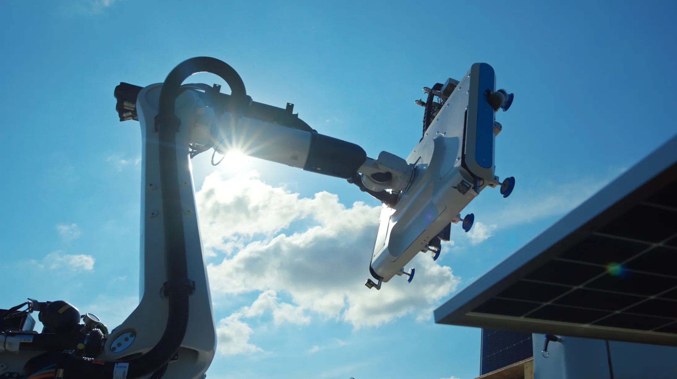 A robotic arm equipped with a large panel is positioned against a bright blue sky with scattered clouds. Sunlight shines through the arm's joints, creating a lens flare. Part of a solar panel is visible in the lower right corner.