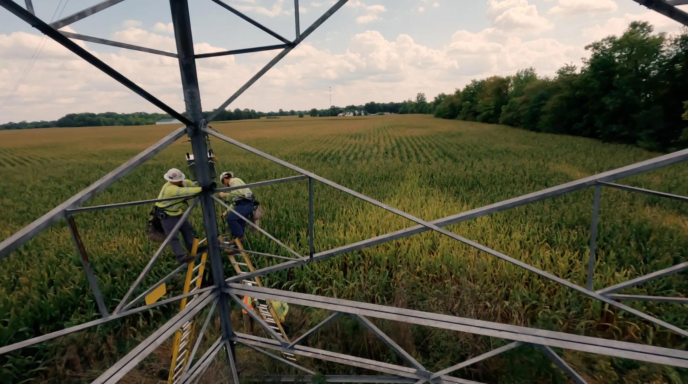 Two workers in safety gear climb a metal tower in a field of corn. The sky is partly cloudy, and trees line the field's edge in the background.