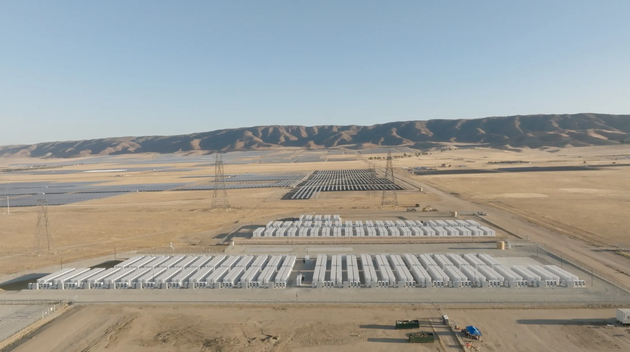 Aerial view of a large solar farm with rows of solar panels and battery storage units in a dry, open landscape. Power lines and distant hills are visible in the background under a clear blue sky.