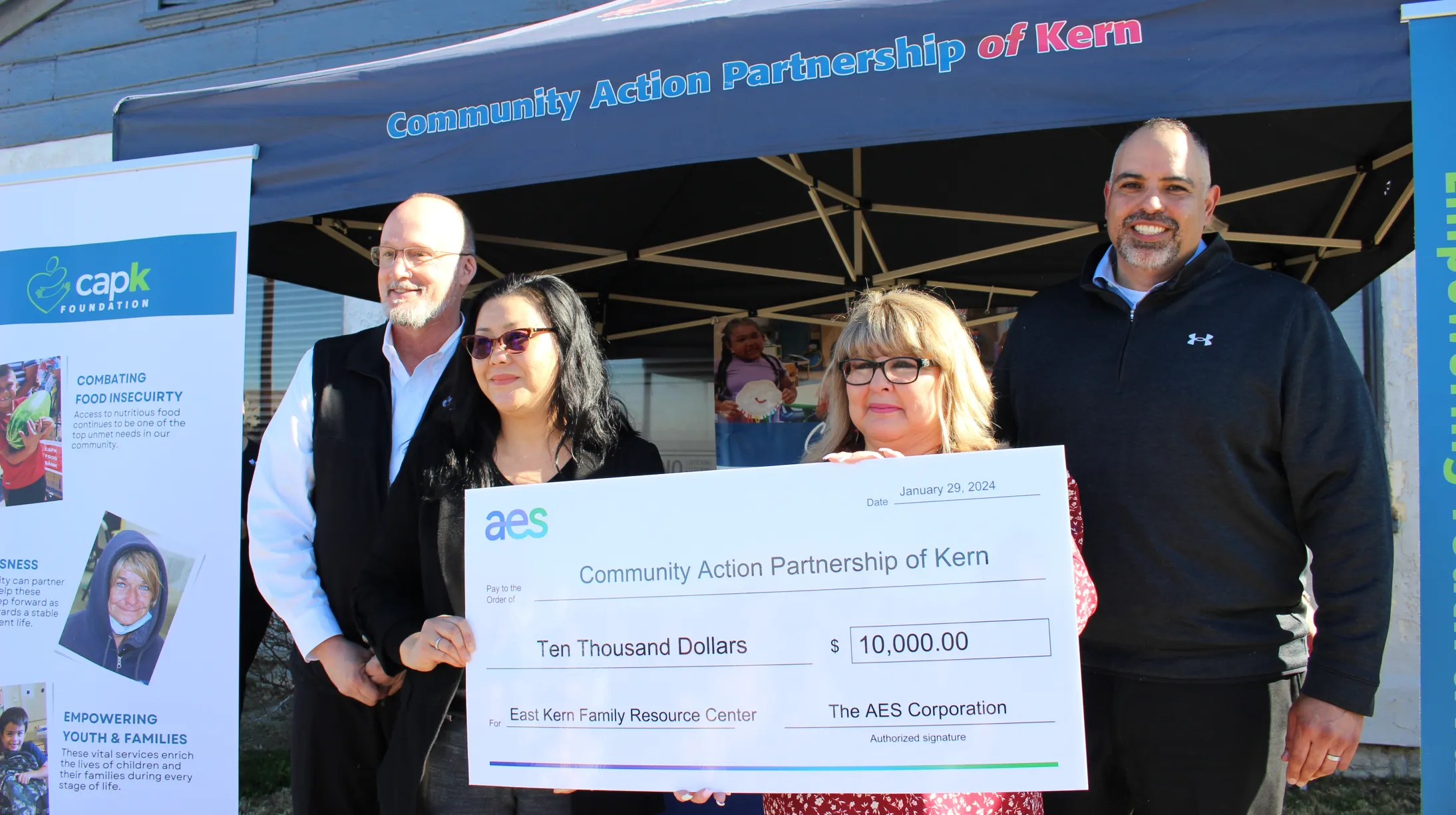 Four people stand in front of a tent labeled 'Community Action Partnership of Kern.' They are holding a large ceremonial check for $10,000 from AES Corporation to the Community Action Partnership of Kern, dated January 29, 2024. A banner and informational posters about food insecurity and community support are visible in the background.