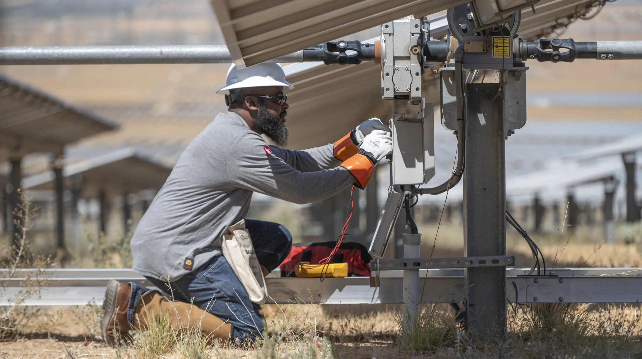 A technician wearing a hard hat and gloves kneels under a solar panel, working on electrical equipment. The technician is using tools and has a tool bag nearby. Rows of solar panels are visible in the background, set in a dry, grassy field.