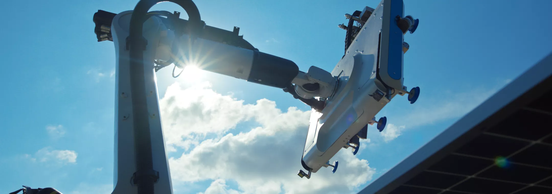 A robotic arm with a tool attachment is positioned against a bright blue sky with scattered clouds. The sun shines through part of the arm, creating a lens flare effect. A solar panel is visible in the lower right corner.