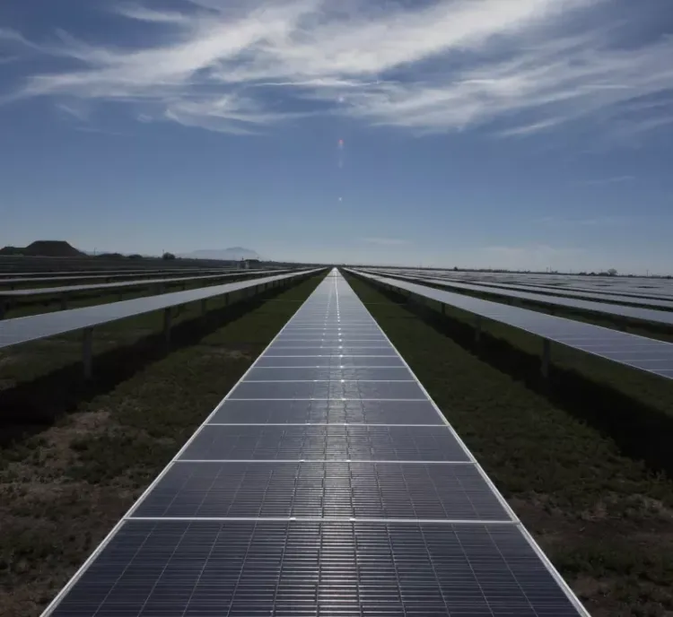 A long row of solar panels stretches into the distance under a blue sky with scattered clouds. The panels are installed on a grassy field, reflecting sunlight. The landscape is flat, and the horizon is visible in the background.