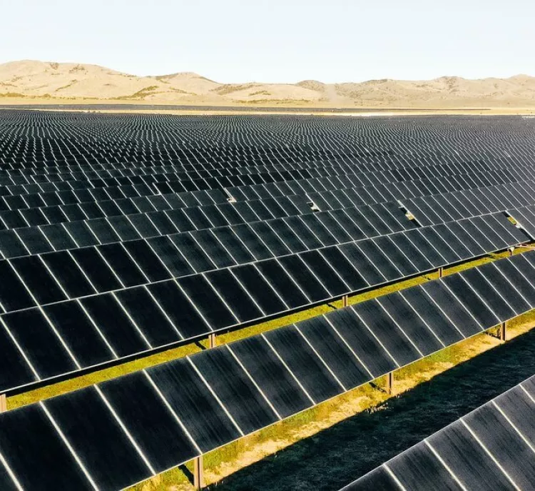 A vast field of solar panels arranged in rows under a clear sky, with distant hills in the background.