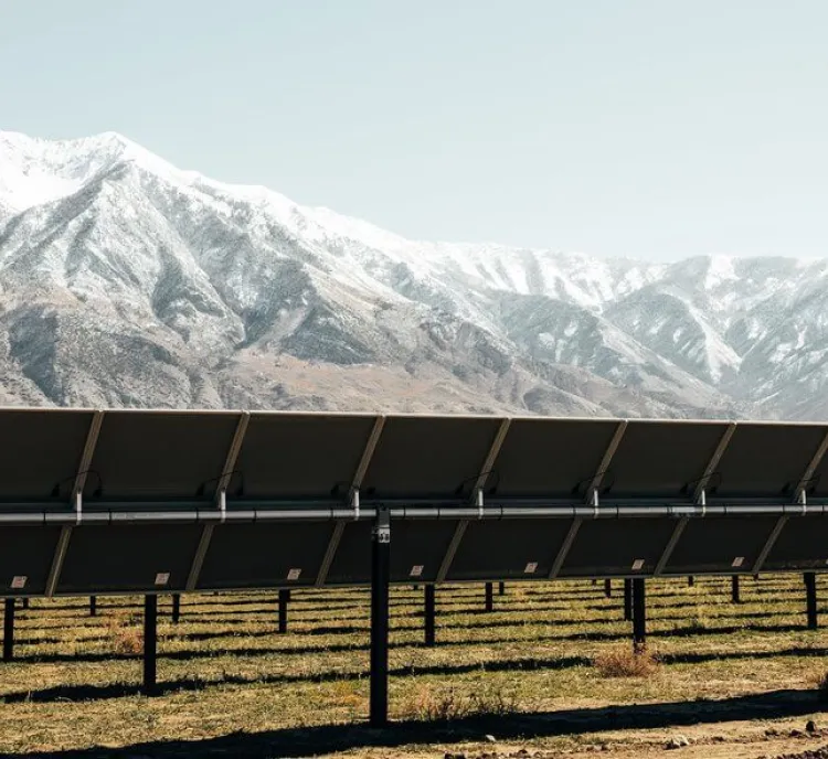 A row of solar panels stands in a field with snow-capped mountains in the background under a clear sky.