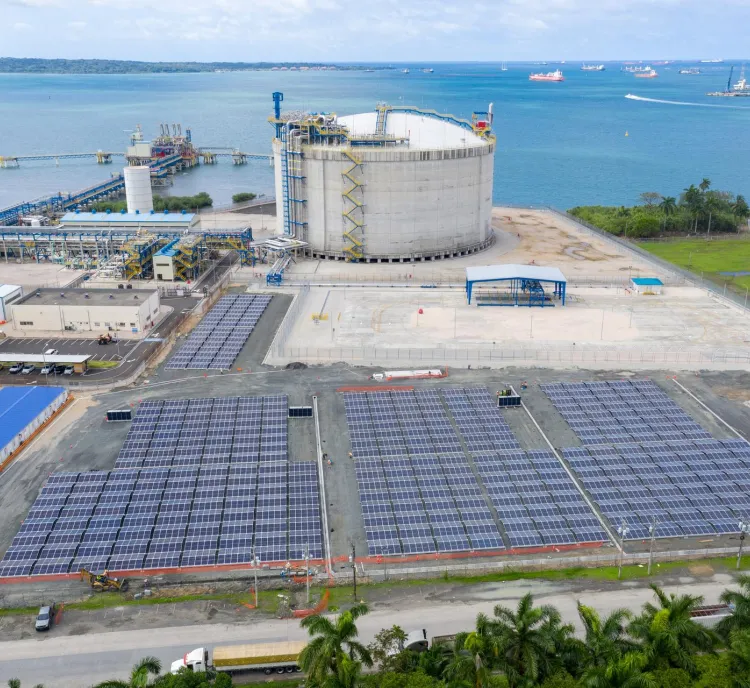 Aerial view of an industrial facility by the sea, featuring a large cylindrical tank, solar panels, and various buildings. The ocean and several ships are visible in the background.