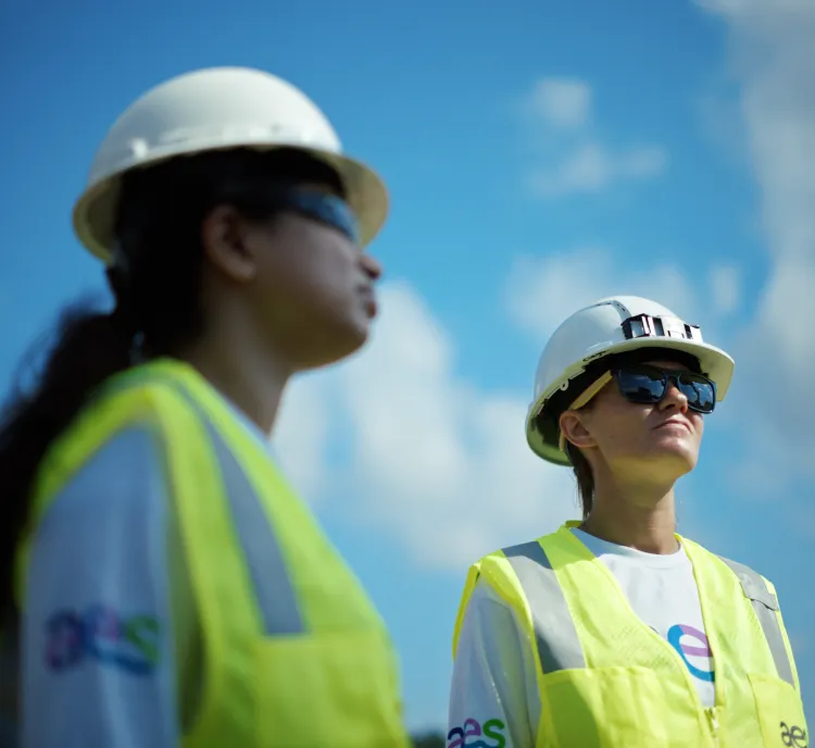 Two people wearing hard hats and reflective vests stand outdoors under a blue sky with clouds. Both are wearing sunglasses and looking upwards.