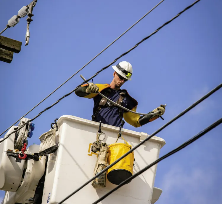 A utility worker wearing a hard hat and safety gear stands in a raised bucket truck, working on power lines against a clear blue sky. The worker is handling equipment with insulated gloves, and a yellow safety bag hangs from the bucket.