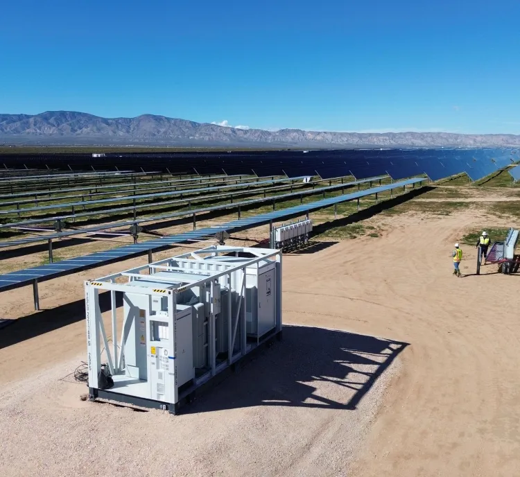 A large solar farm with rows of solar panels extending into the distance under a clear blue sky. In the foreground, there is a white equipment container on a dirt path. Two workers wearing yellow vests are near a set of panels, inspecting or working on them. Mountains are visible on the horizon.