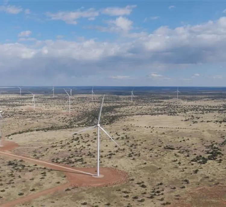 Aerial view of a wind farm in a vast, open landscape under a partly cloudy sky. Several wind turbines are scattered across the terrain, with dirt roads visible among sparse vegetation.