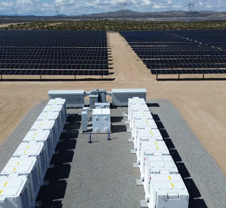 Aerial view of a large solar power plant in a desert landscape. Rows of solar panels stretch into the distance. In the foreground, there are several white electrical units and equipment on a gravel area. Power lines and mountains are visible on the horizon under a partly cloudy sky.