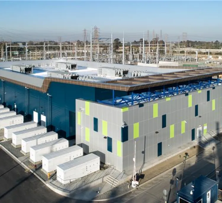 Aerial view of a large industrial building surrounded by electrical equipment and power lines. The building has a modern design with blue and gray panels and green accents. Rows of white containers are aligned along one side. The surrounding area includes roads and additional industrial structures under a clear blue sky.