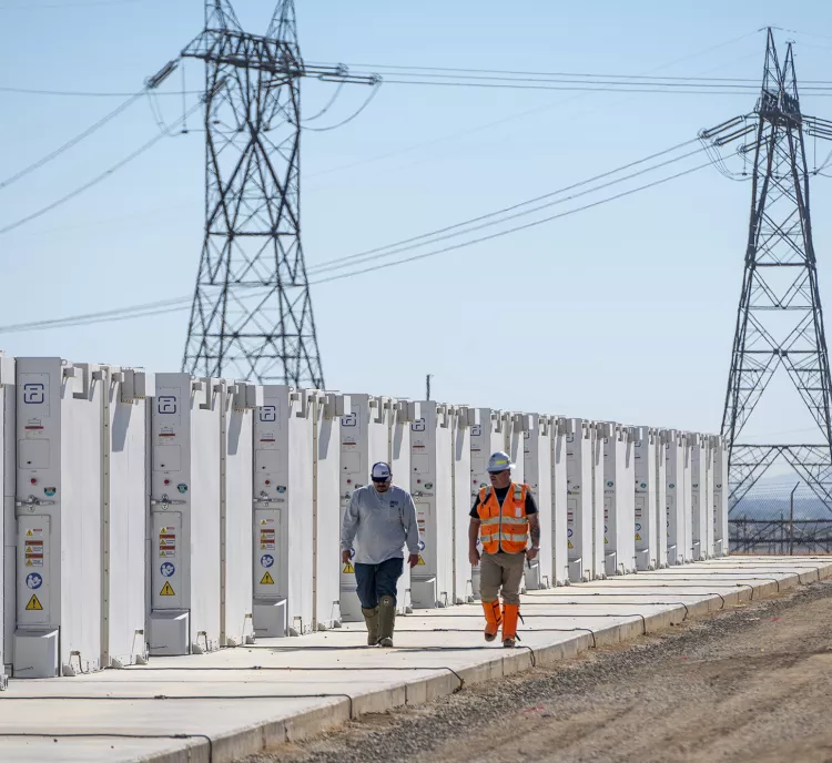 Two workers in safety gear walk along a row of large white battery storage units at a solar facility. High-voltage power lines and towers are visible in the background under a clear blue sky.