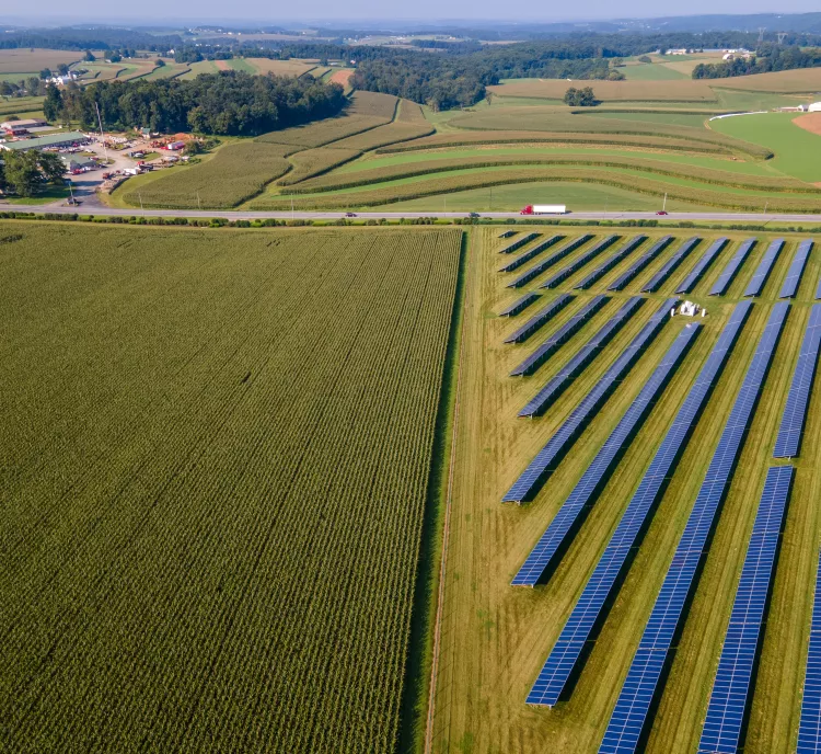 Aerial view of a large solar farm with rows of solar panels on the right side, adjacent to vast green agricultural fields on the left. In the background, a small rural community with buildings and roads is visible, surrounded by rolling hills and more farmland.