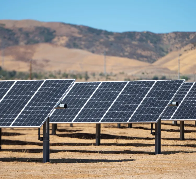 Rows of solar panels installed in a dry, open field under a clear blue sky, with rolling hills in the background.