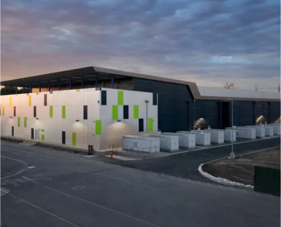 A large industrial building with a modern design, featuring a facade with green, black, and white panels. The building is surrounded by a paved area and has several white utility boxes lined up along its side. The sky is overcast with a hint of sunset light.