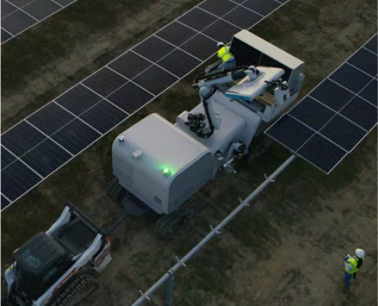 Aerial view of a solar farm with rows of solar panels. A large machine with a robotic arm is stationed between the panels, attended by workers in safety gear. A smaller vehicle is parked nearby. The setting is outdoors on a dirt surface.