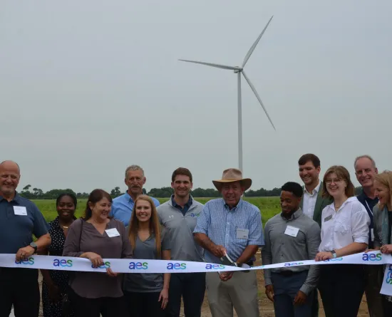 A group of people standing outdoors in front of a wind turbine, smiling and holding a ribbon with the AES logo. One person is cutting the ribbon, marking a ceremonial event. The background shows a grassy field and a cloudy sky.