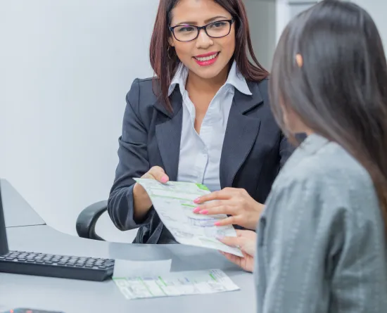 A woman in a business suit and glasses is sitting at a desk, smiling and holding documents while talking to another person across the table. A keyboard and additional papers are visible on the desk.
