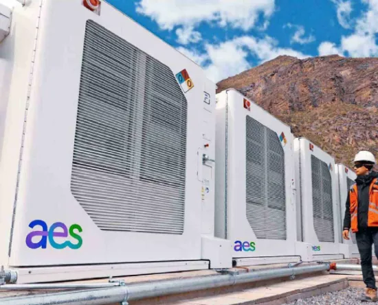 A person wearing a hard hat and orange safety vest walks past large white industrial units with the AES logo. The units are aligned in a row with a mountainous landscape and cloudy sky in the background.