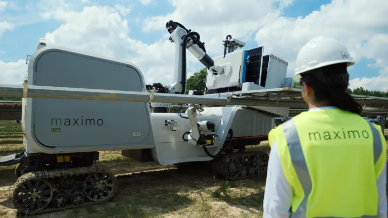A person wearing a yellow safety vest and white hard hat stands in front of a large robotic machine labeled 'maximo aes' on a tracked base. The machine is equipped with a robotic arm and solar panels, set in an outdoor environment with a partly cloudy sky.