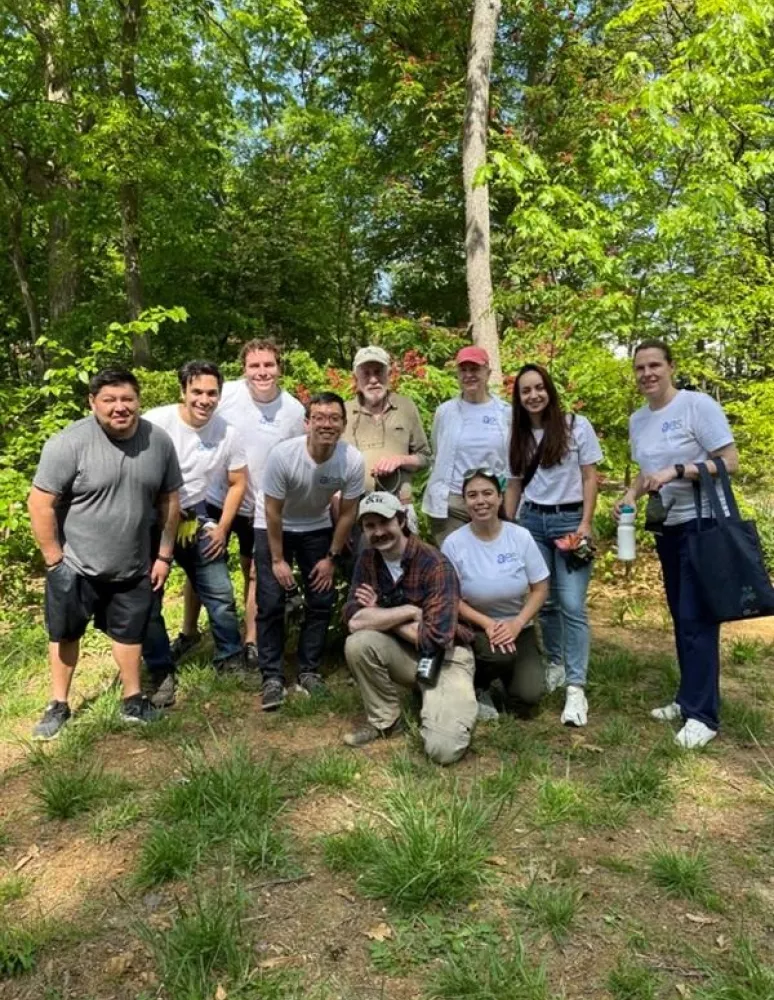 A group of eleven people, some wearing white shirts with a logo, pose together outdoors in a wooded area. They are standing on grass with trees in the background. One person is crouching in front, wearing a plaid shirt and cap. The group appears to be enjoying the sunny day.