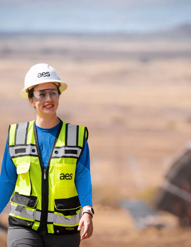 A person wearing a white hard hat with 'aes' logo, safety glasses, and a high-visibility vest, walking outdoors near solar panels in a desert landscape.