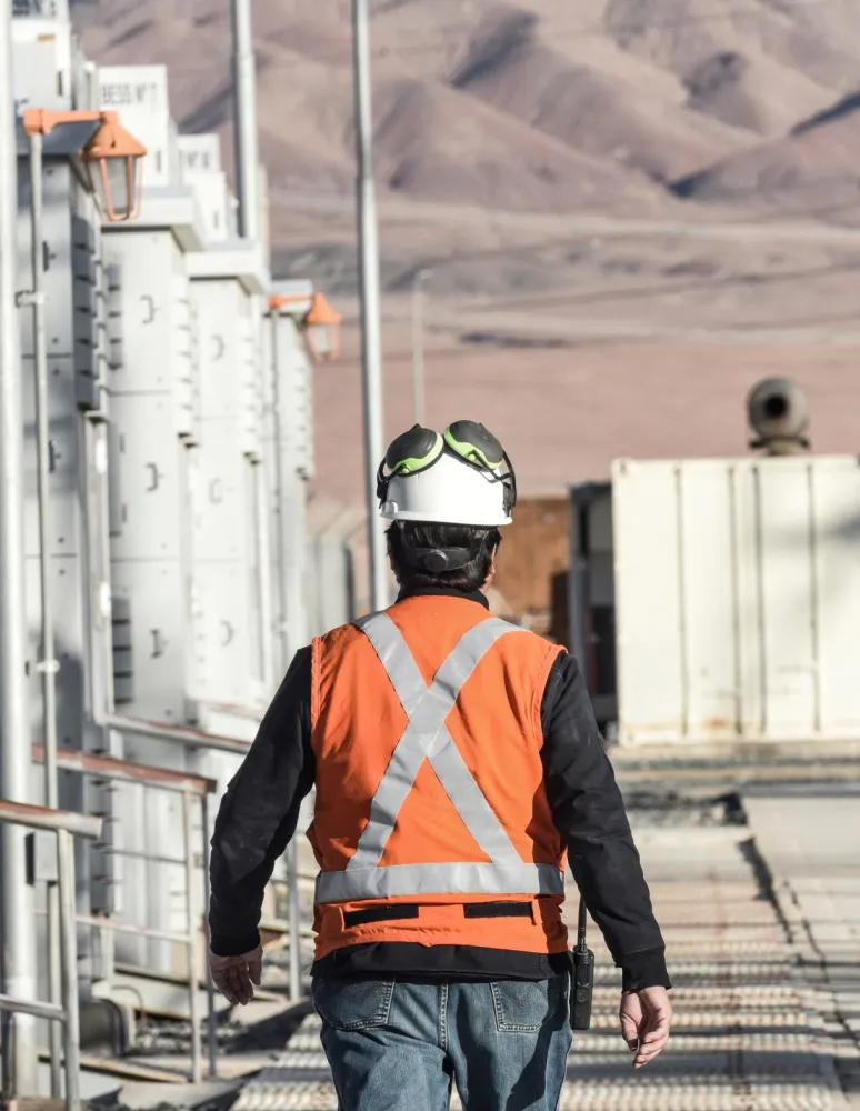 A person wearing an orange safety vest and white helmet walks between industrial equipment in a desert landscape. The background shows arid mountains under a clear sky.