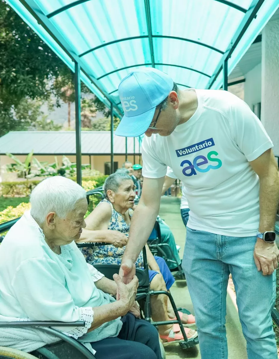 A volunteer wearing an AES shirt and cap is bending down to shake hands with an elderly woman in a wheelchair. Another elderly woman is seated nearby under a covered walkway with greenery around.
