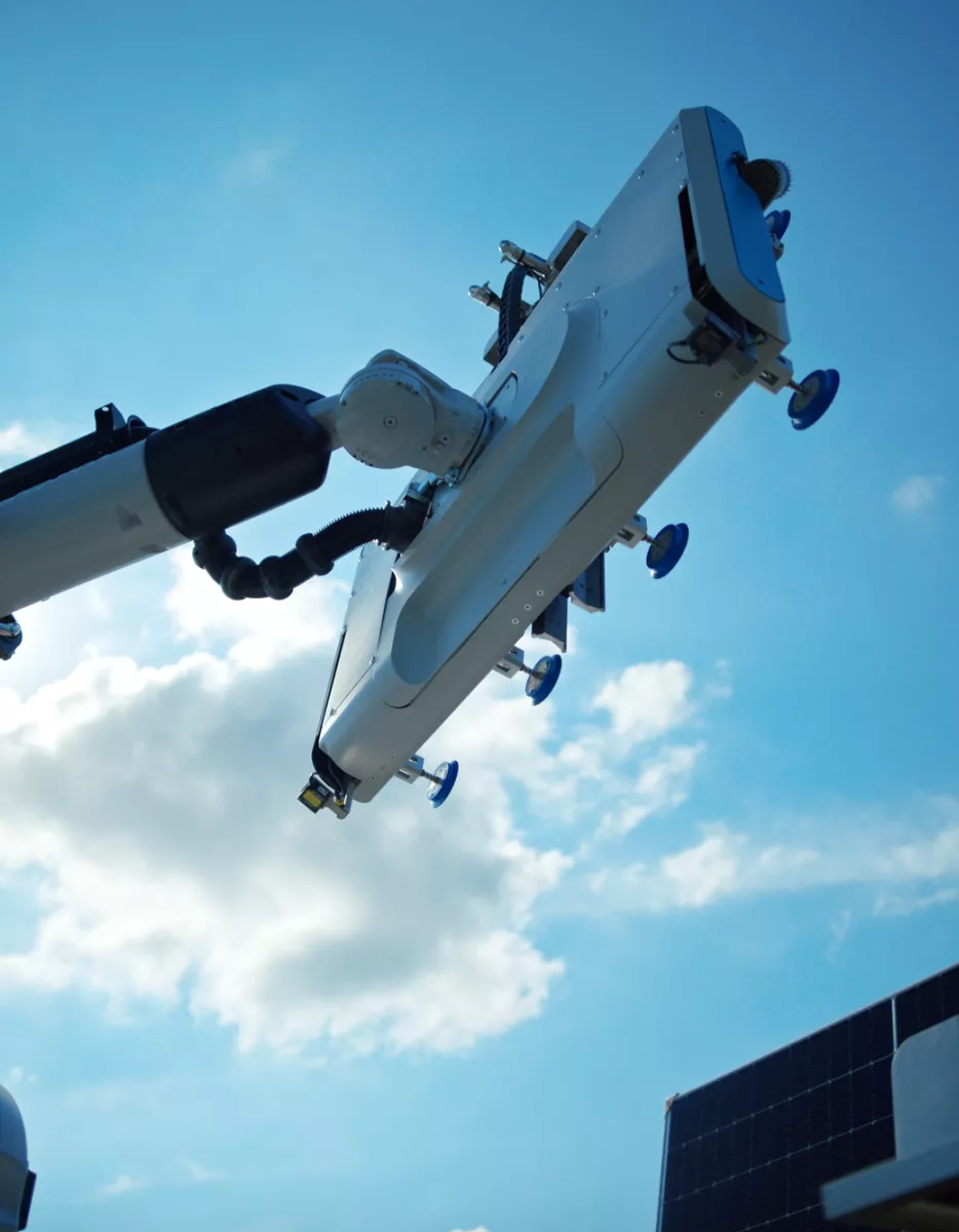 A robotic arm with a large rectangular attachment reaches towards the sky, set against a backdrop of a clear blue sky with scattered clouds. In the lower right corner, part of a solar panel is visible.
