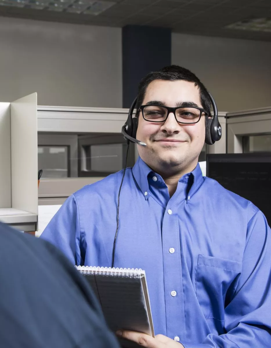 A man in a blue shirt and glasses wearing a headset stands in an office with cubicles, holding a notebook and smiling at a woman with her back to the camera.