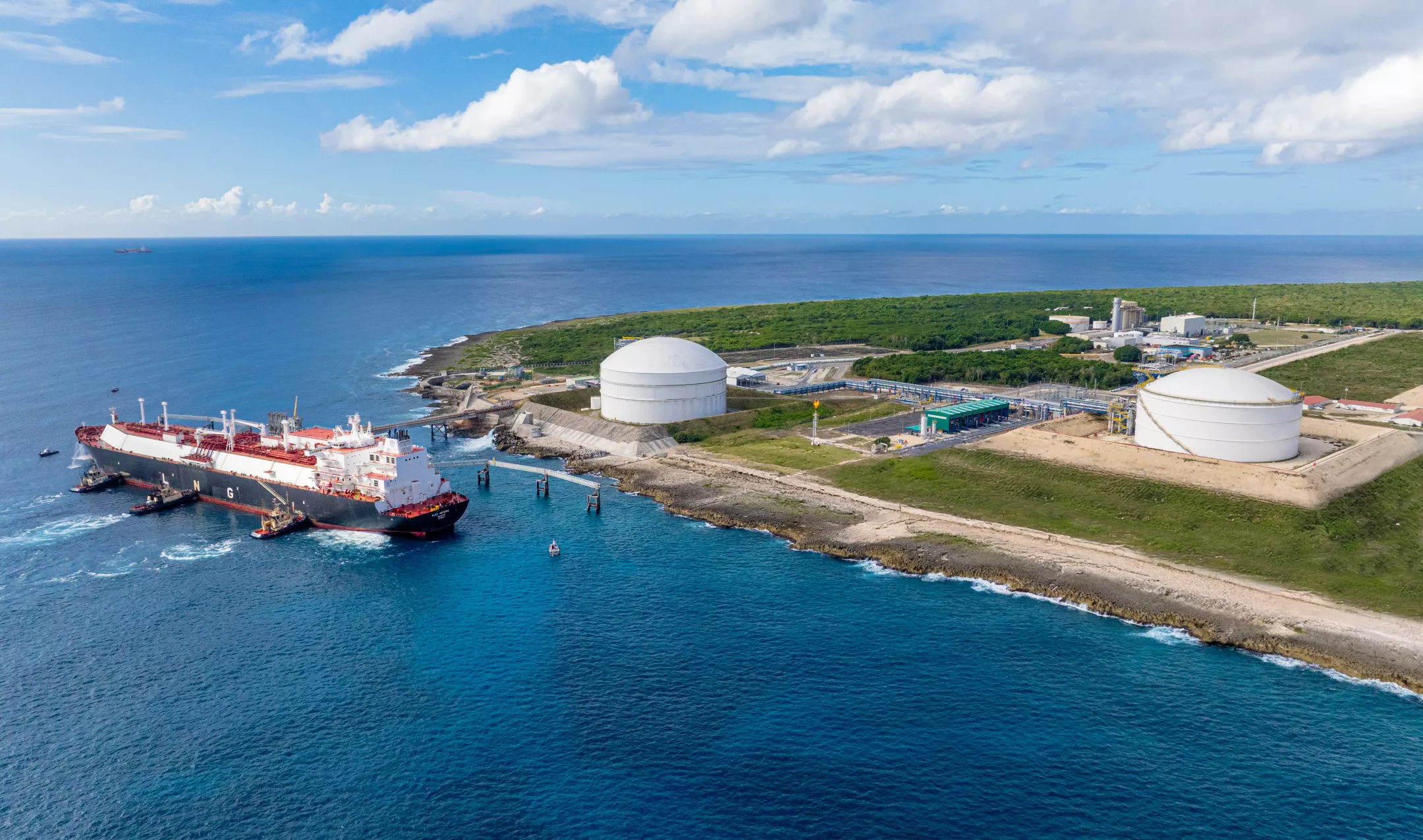 Aerial view of a large tanker ship docked at a coastal LNG terminal with two large white storage tanks. The terminal is situated on a green, grassy landscape next to the ocean under a partly cloudy sky.