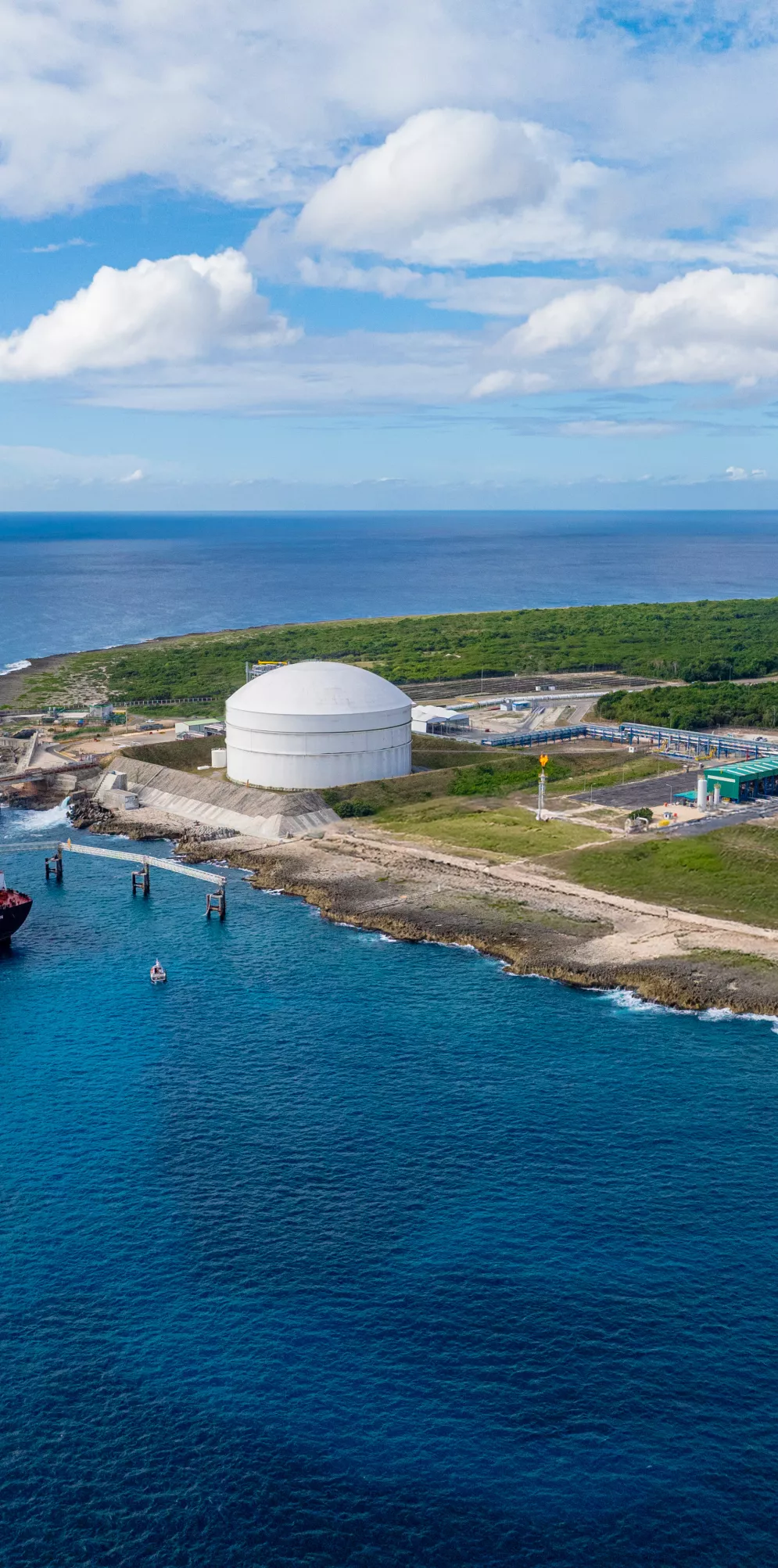 Aerial view of a large tanker ship docked at a coastal LNG terminal with two large white storage tanks. The terminal is situated on a green, grassy landscape next to the ocean under a partly cloudy sky.