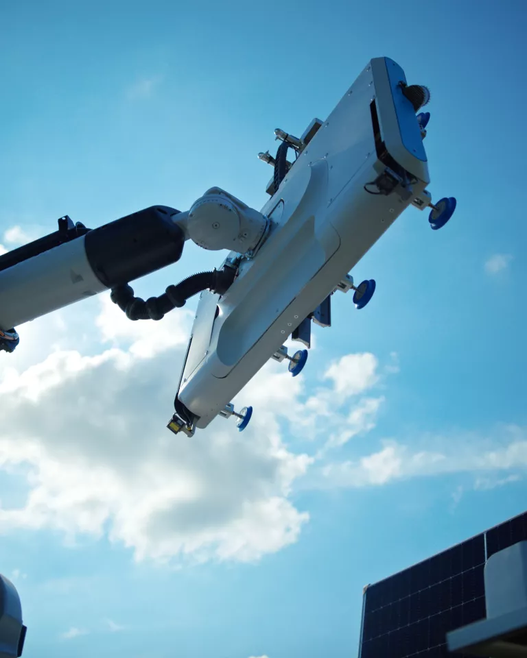 A robotic arm with a flat, rectangular attachment extends upward against a bright blue sky with scattered clouds. The arm is part of a larger structure visible in the lower right corner, resembling solar panels.