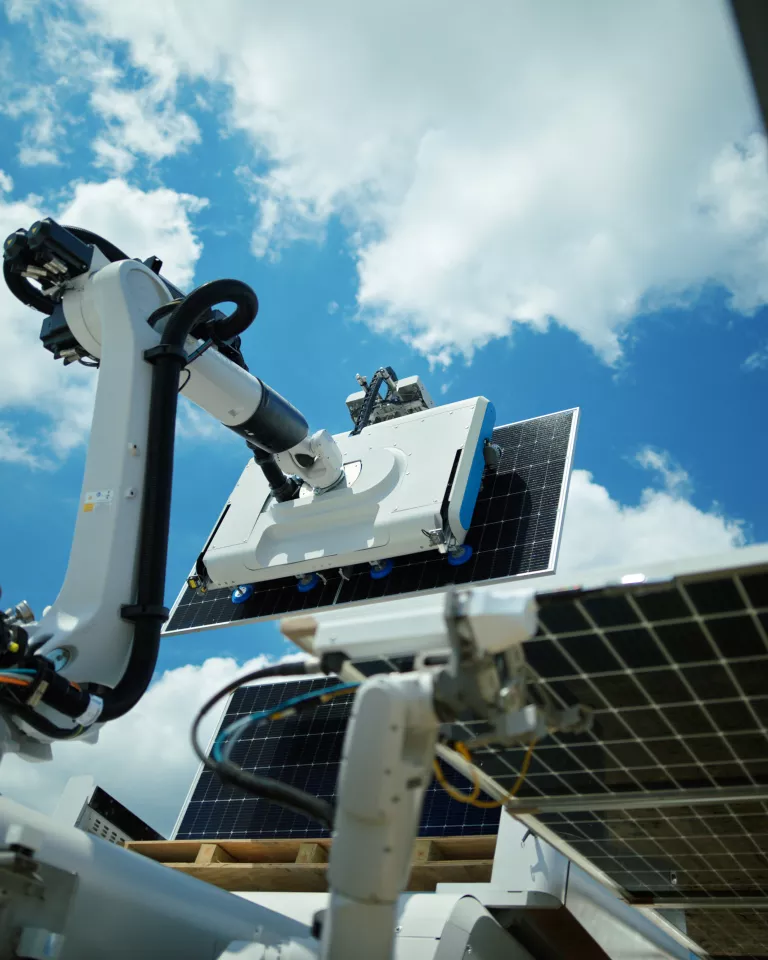 A robotic arm with a cleaning device is positioned over a solar panel under a bright blue sky with scattered clouds.