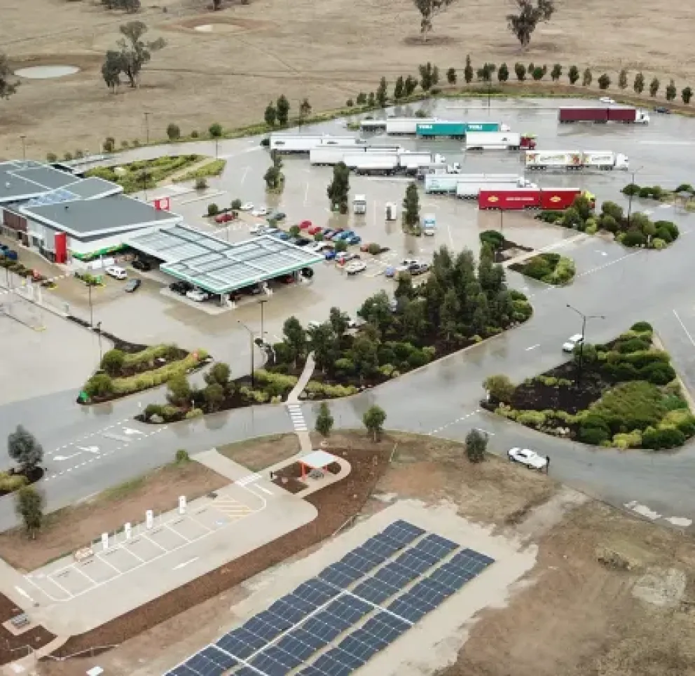 Aerial view of a large truck stop with multiple parked trucks, a convenience store, and fuel pumps. The area is surrounded by open land with scattered trees and features solar panels in the foreground.