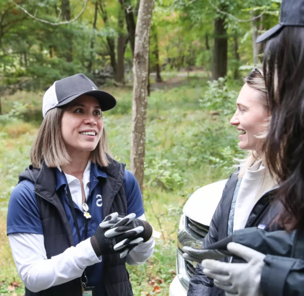 Three people wearing outdoor gear converse in a forested area. The person on the left smiles and gestures with gloved hands. Trees and greenery are visible in the background.