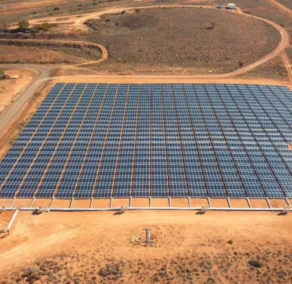 Aerial view of a large solar panel farm in a desert landscape. The solar panels are arranged in a grid pattern on sandy terrain, surrounded by dirt roads. A small white building is visible in the bottom left corner.