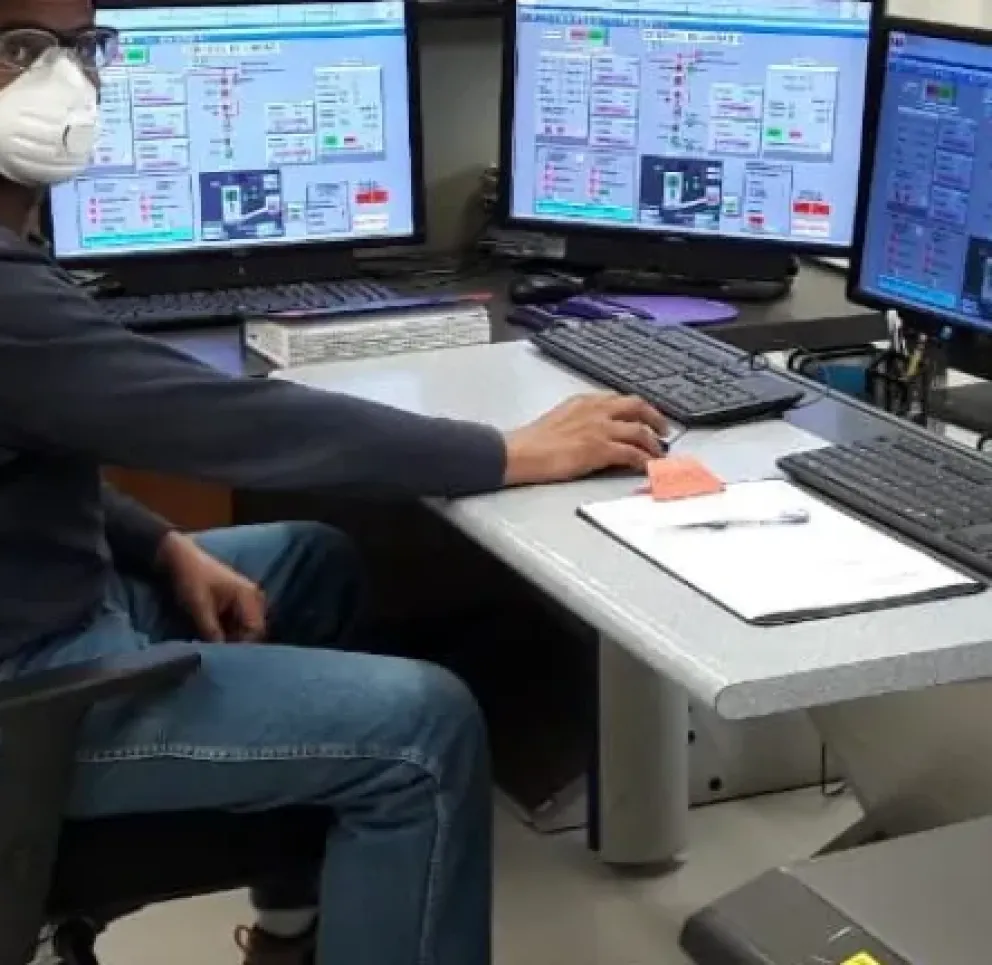 A person wearing a mask and hard hat sits at a control desk with multiple computer monitors displaying complex data and graphs.
