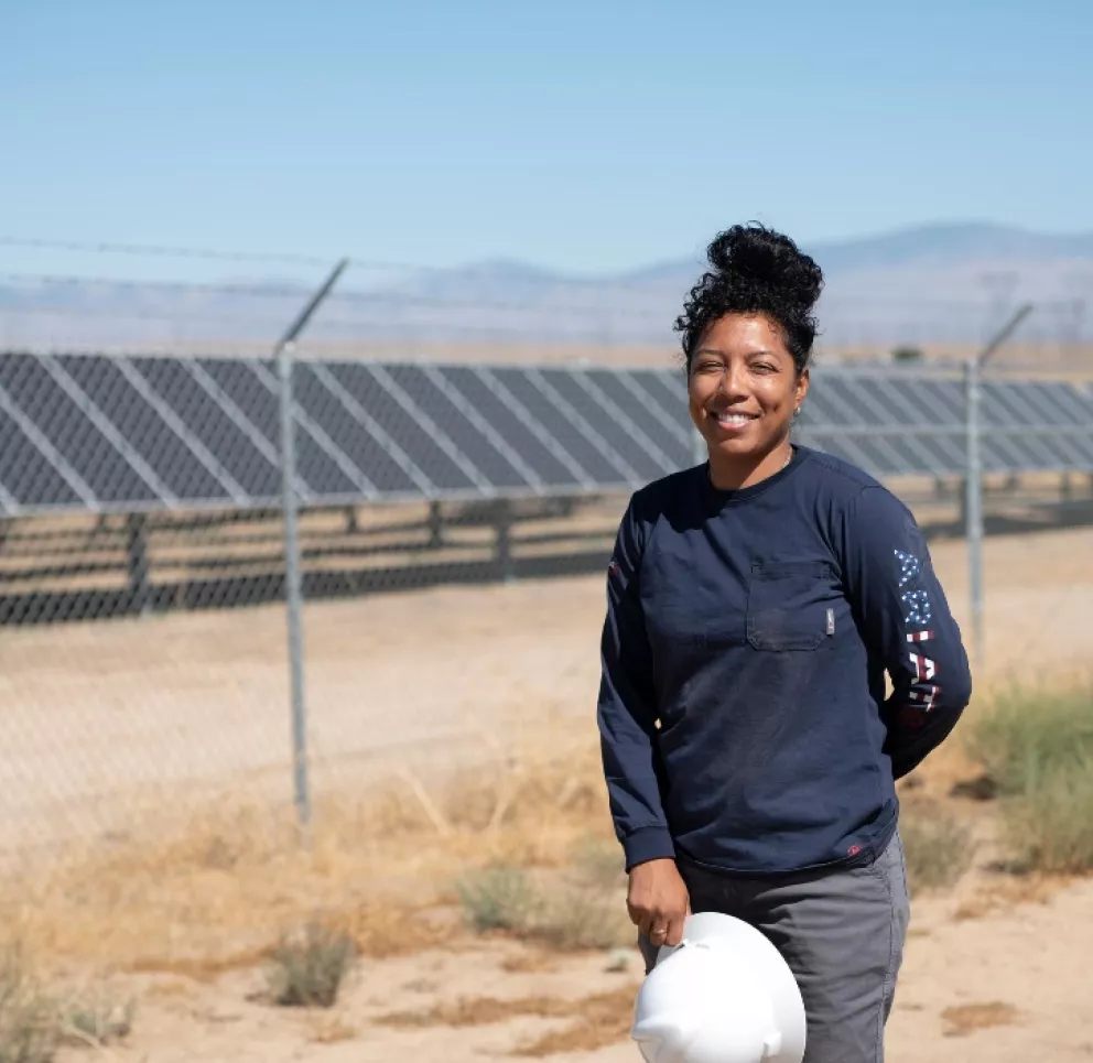 A person in a dark long-sleeve shirt stands holding a white hard hat, smiling in front of a fenced area with rows of solar panels under a clear blue sky. Dry grass and mountains are visible in the background.